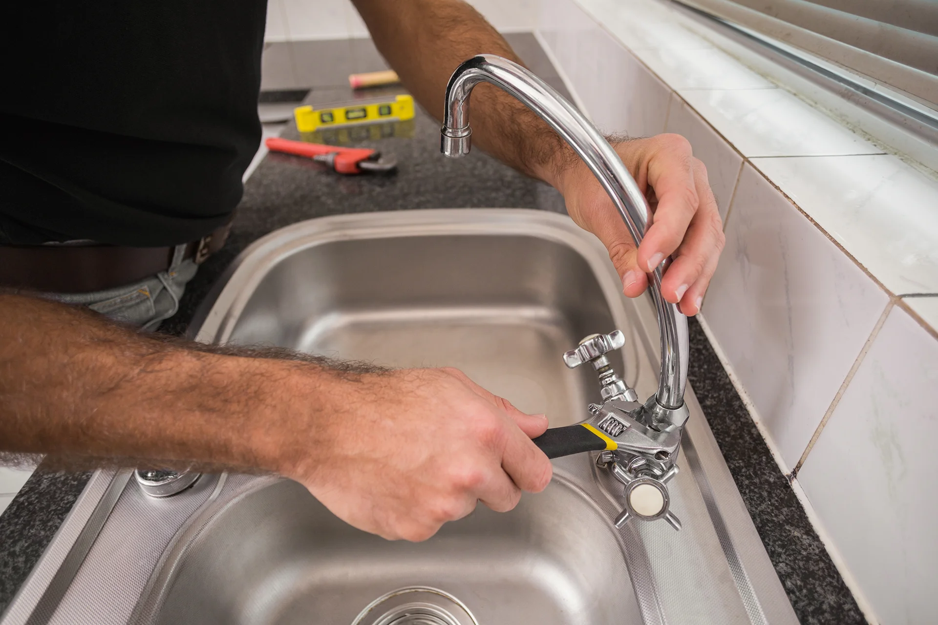 A plumber tightening a chrome faucet with an adjustable wrench over a stainless steel kitchen sink.