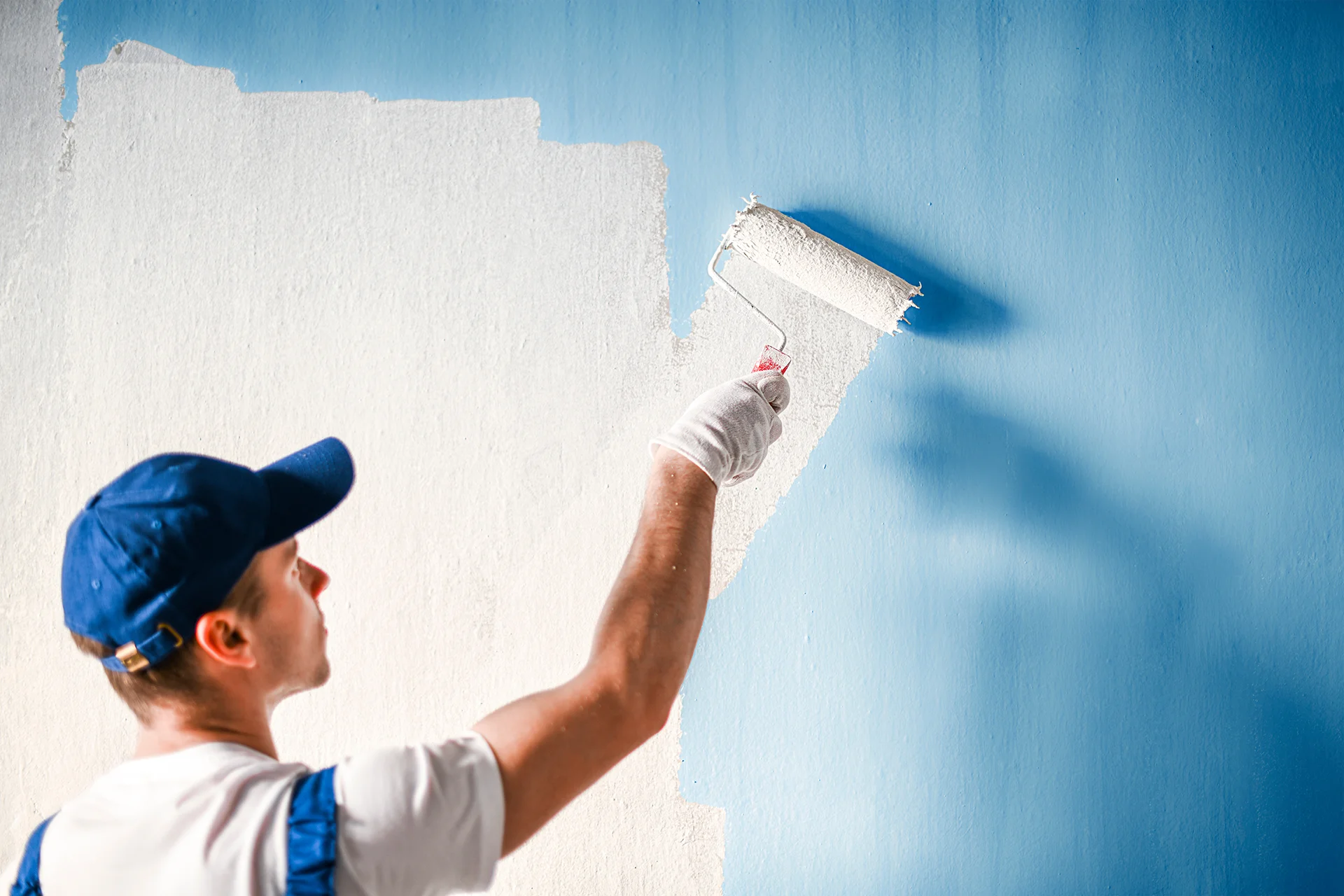 A worker wearing a blue cap and overalls using a roller to paint a wall white over a blue surface.