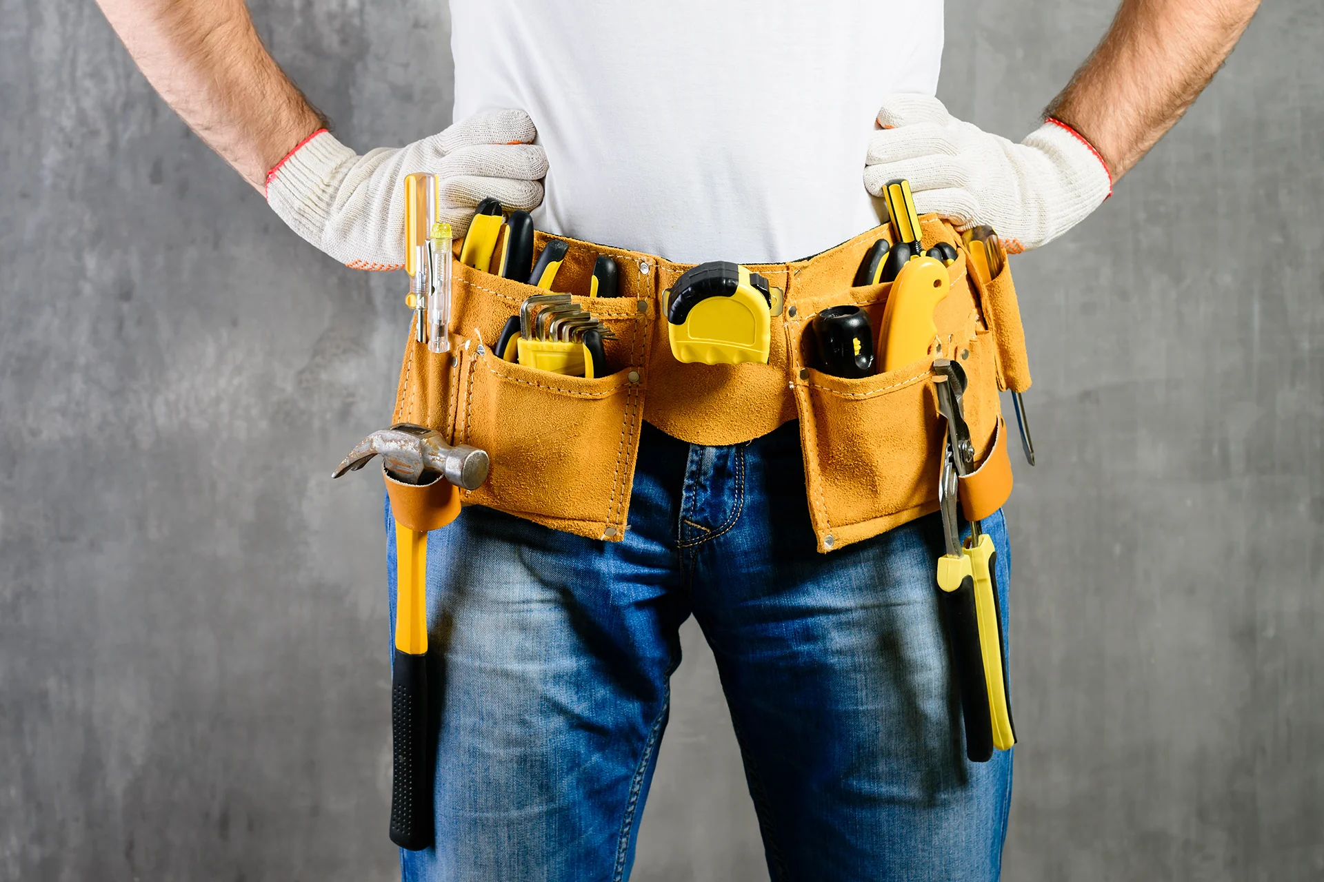 A worker wearing a tan leather tool belt filled with tools and gloves over a white t-shirt and jeans.